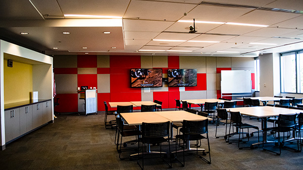 Global room set up as 6 squares of tables with chairs facing wall with monitors.