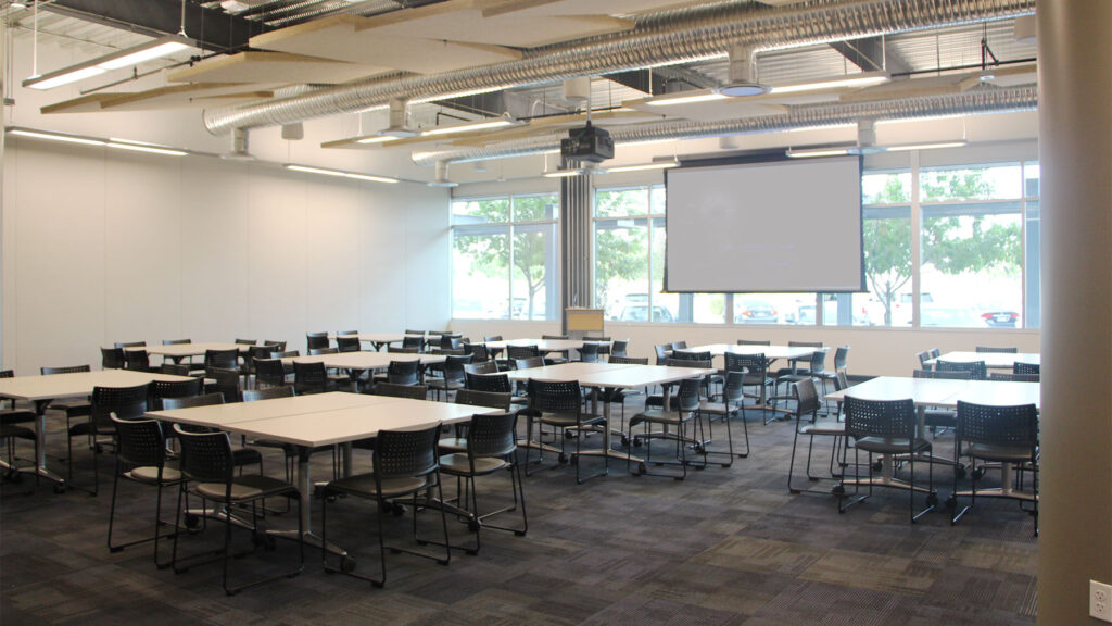 Synergy II room set up as square tables seating 8, with 10 sets, from back of room. Screen lowered down against windows.